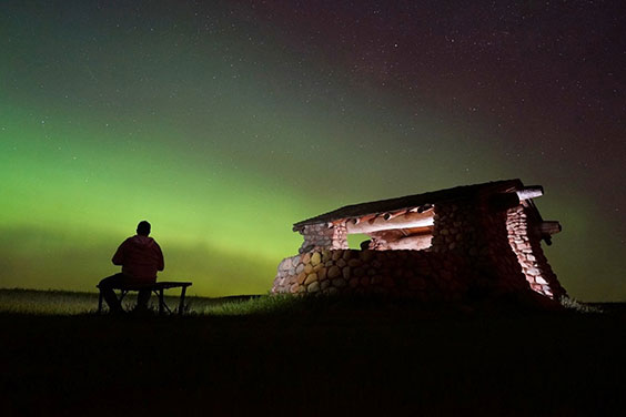 A night photo of a stone shelter that's lit from the inside. Next to the shelter is the silhouette of a man sitting on a bench. The sky has streaks of green from the Northern Lights.