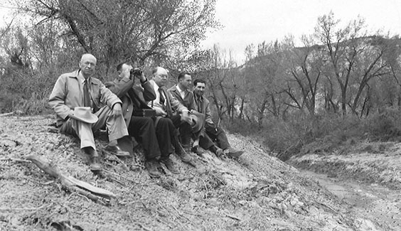 Five men dressed in pants, button up shirts, ties, and jackets sit on a hill with trees in the background