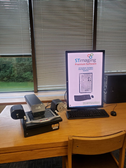 A computer monitor with microfilm processing equipment next to it sit on a wooden table with windows with drawn blinds in the background