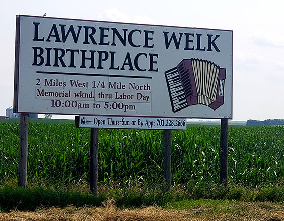 Large white road sign that reads Lawrence Welk Birthplace - 2 miles west 1/4 mile north - Memorial wknd. thru Labor Day - 10:00am to 5:00pm. There is also a graphic of an accordion on the sign.