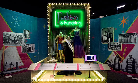 The entrance for an exhibit has the title Fashion & Function: North Dakota in pink and green neon lights. There are 3 mannequins below the sign. One is wearing a red and blue wonder woman costume. One is wearing a dark colored pageant gown with a Miss America sash. The third is wearing a burple ballgown with silver gems ont the top.