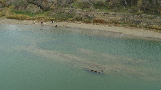 A shipwreck can be seen just below the water while a few people stand along the shoreline