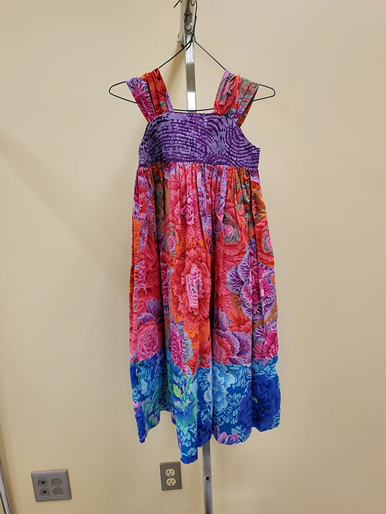 A red dress with a section of purple across the bust and blue across the bottom with purple flowers throughout hangs from a metal hanger