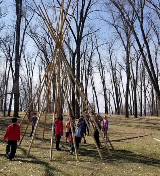 One adult and many students stand inside and around the wooden poles of a tipi that's setup without the covering.