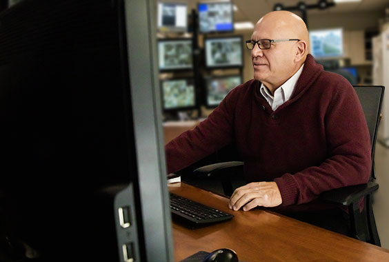 male security guard in maroon sweater and glasses in control room with monitors 