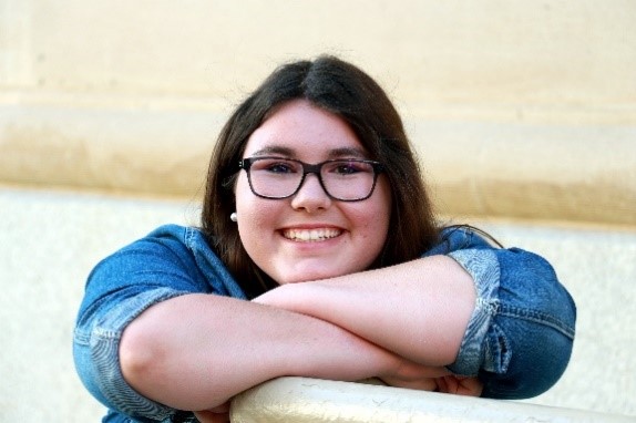 A young girl with dark hair, glases, and a blue shirt poses with her arms crossed and chin resting on them