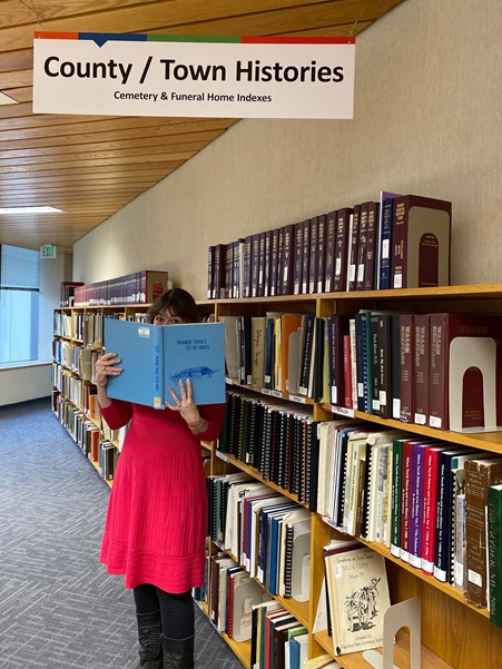 A woman in a red dress and black leggings stands in front of wall lined with 5 rows of books and is holding a light blue book with dark blue writing in front of her.