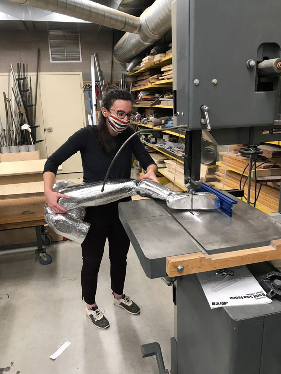 A woman with black pants and shirt, black glasses, darnk har, and a red, white, and blue striped face mask stands holding a tinfoil wrapped mannequin leg on a table saw, about to cut part of the foot off