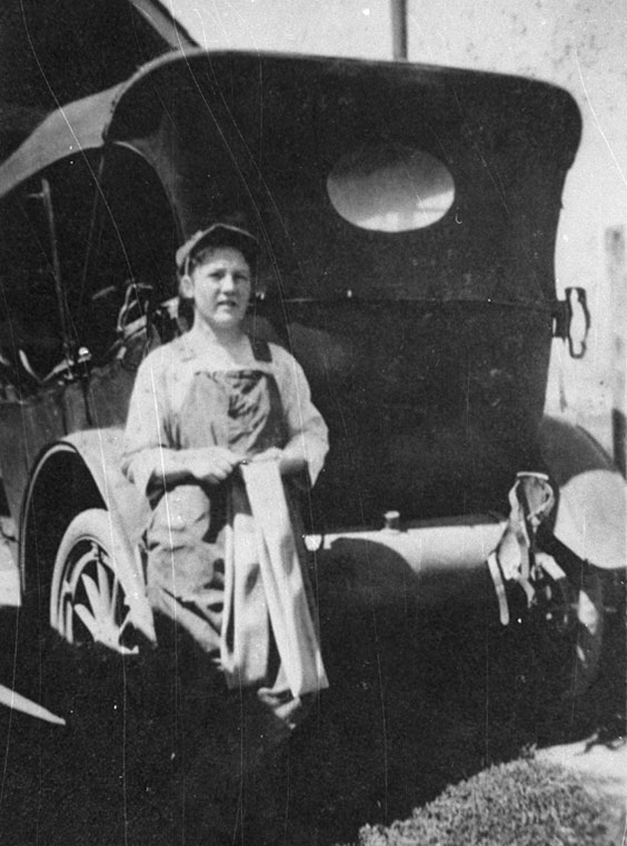 A woman stands in front of an old automobile patching the inner tube of a tire