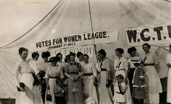 A group of women stand in front of a tent that reads FOVES FOR WOMEN LEAGUE (NOVEMBER 3RD 1914)