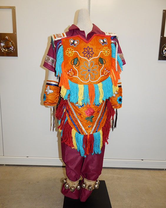 Maroon collared short sleeved shirt and pants with a bright orange chest plate emboridered with green, blue. red, yellow, and white flowers and butterflies. There are teal, orange and yellow strings of beads or yarn coming out of the sides and bottom of the chest plate. There is also an orange piece around the waist that hangs down to the knee area as well as wrist cuffs that look similar to the chest plate. Around the ankles are tan cuffs with silver bells attached to them.