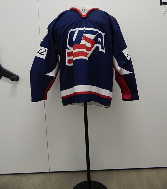 A blue hockey jersey with red around the neck, white patches on the arms, white and red down the inside of the arms, and a thick white and thinner red band around the bottom of the jersey. In the middle of the jersey is a logo that says USA in blue with a white outline. The bottom half of the S is red and turns into a waving flag. The inside of the A is a white star.