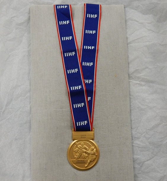 A gold medal with a goalie and hockey player skating with their stick up in the air as if trying to score a goal. The part that goes around the neck is blue with a thin white and thicker red borders and reads IIHF repeatedly in white lettering.