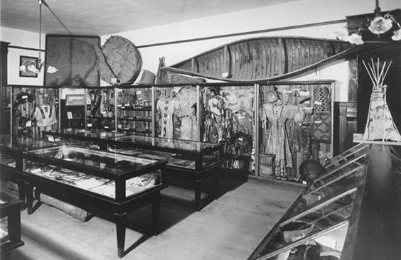 An exhibit with display cases lining the walls and glass tables in the middle. Many artifacts are displayed. Above the cases is a canoe and bull boat and other artifacts.