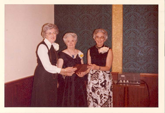 Three older women in dresses stand holding an award. They each wear a white corsage on their left side.