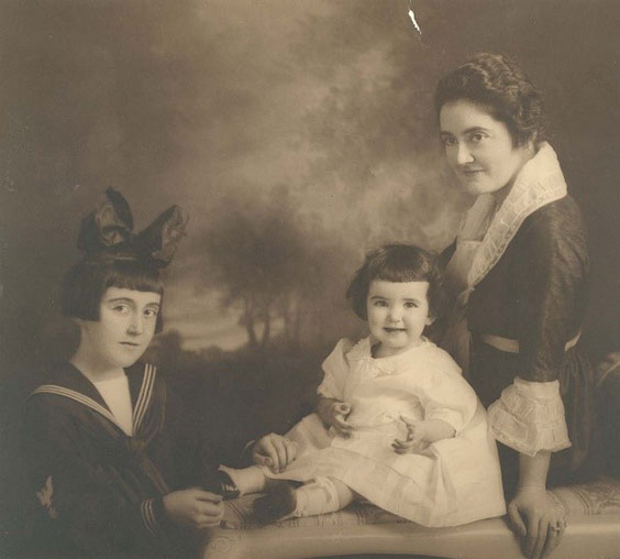 A mother and two daughters pose for a picture. The mother wears a darc colored dress with white lace around the neck and cuffs and a white belt around the waist. The youngest child wears a  white puffy dress. The other child wears a dark colored dress with three white lines around the collar and wrists. She also wears a large bow in her hair.