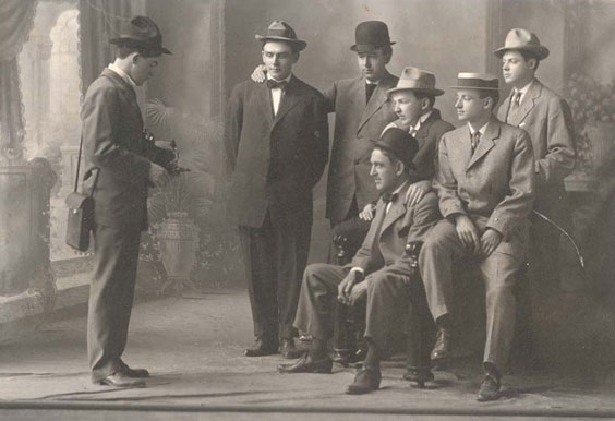 6 men sit and stand together while another man stands across from them looking down at something in his hands. The men wear sack suits featuring a boxy cut with a higher neck line and shorter lapels. They are all also wearing hats.