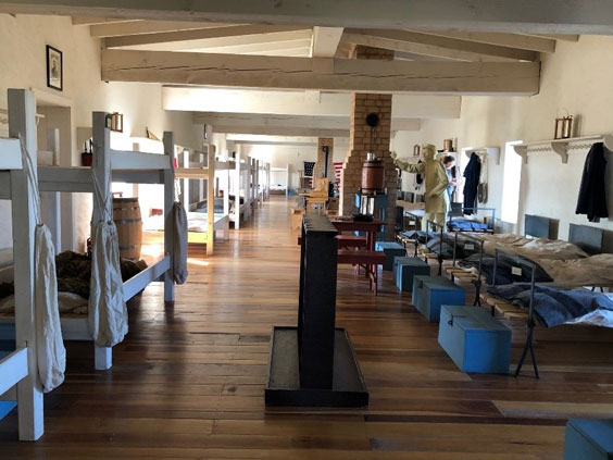 The interior of barracks with white wooden bunk beads and black framed cots with blue trinks at the foot of them.