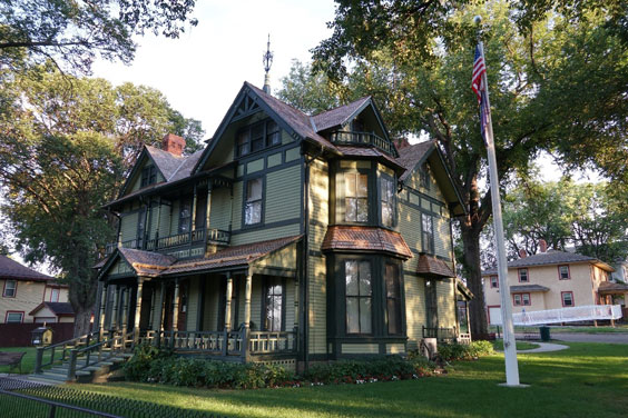 The exterior of an old, large, light green house with dark green trim and brown shingles. There is a frong porch on the house.
