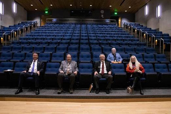 A view looking from the stage of an auditorium out towards the crowd. Three men and two women sit among the blue cushy chairs.