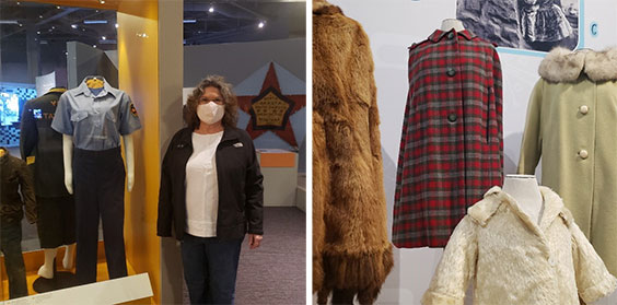 A woman wearing dark pants, white shirt, black jacket, and white face mask stands next to an exhibit case displaying a uniform with dark pants and a short sleeved light blue button up shirt.