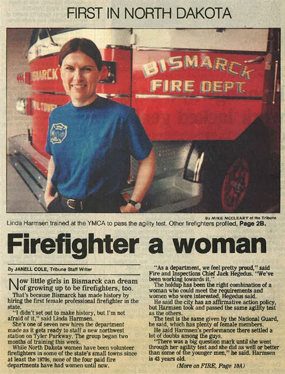 A woman wearing a blue tshirt and dark pants with her hair tied back stands in front of a red fire truck that reads Bismarck Fire Dept. The headline of the news article below the image reads Firefighter a woman.