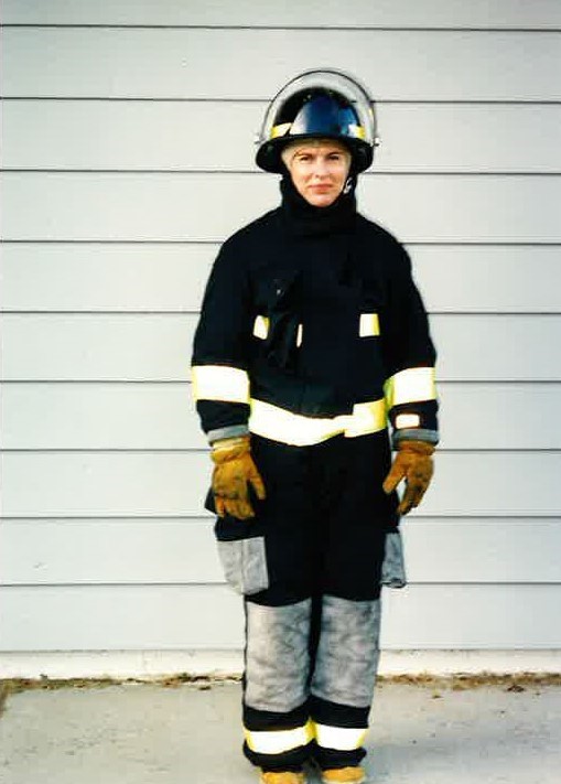 A woman stand in full firefighter turnout gear that is black with gray knees and pockets and yellow reflective trim.