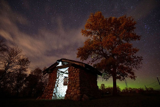 Night scene of a stone shelter that's lit inside. Trees can be seen around the shelter as well as a green glow at the horizon from the northern lights and many starts in the sky and a few clouds.