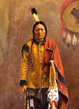 A Native American man stands wearing a cape over one shoulder, feather behind his head, tan long sleeved shirt with darker cuffs and shoulders. His long, dark hair is in two braids.