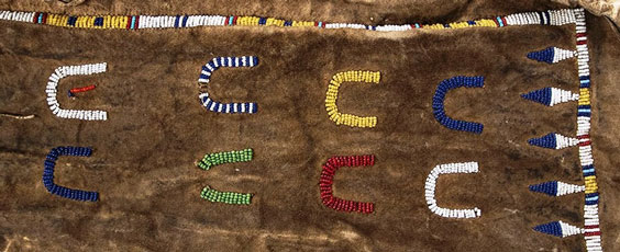 Detailed view of a pipe bag with white and yellow beaded trim with white and blue diamonds along the bottom above the trim. White, blue, green, white and blue, yellow, red, white, and blue beaded horseshoes decorate the bag. 