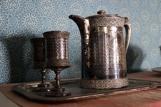 Wilver pitcher and two cups. Each have a floral pattern border around the top and bottom and square designs between the borders. The wallpaper in the background is blue with tan and darker blue designs in circles.