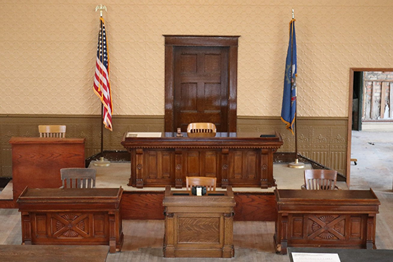 A judge's wooden bench sits rear center. To the left of it is a smaller desk. In front of it are three desks with the middle one being smaller and lighter in color. The American flag and North Dakota flag stand on either side of the judge's desk.