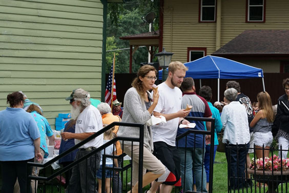 many people are gathered outside for an event. Some are eating ice cream cones. A blue canopy is set up between a green building and a yellow house.