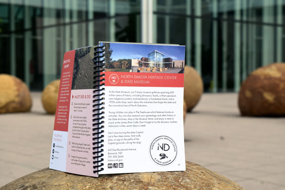 A spiral bound book open to a page reading North Dakota Heritage Center & State Museum with a stamp on it sits on top of a round rock with a glass building in the background