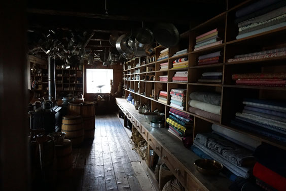 A room with wooden shelves lining one wall. On the shelves are folded, colorful fabric