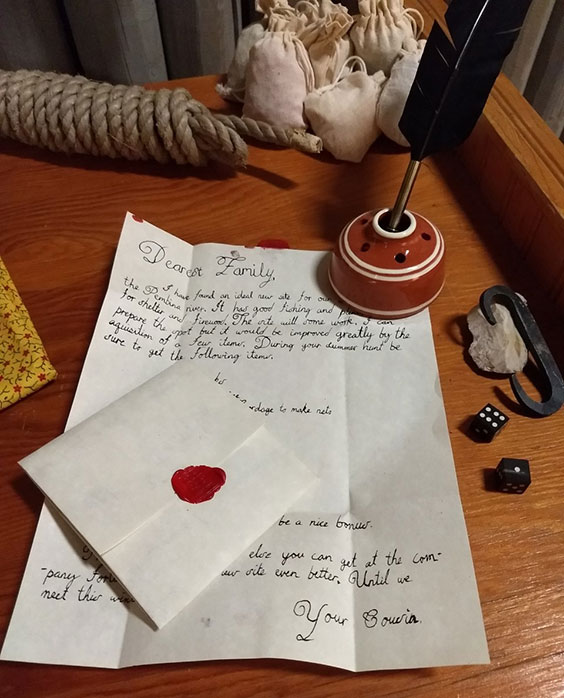 A hand inked letter, envelope with wax seal, quill and ink, rope, dice, and other items sit atop a wooden table