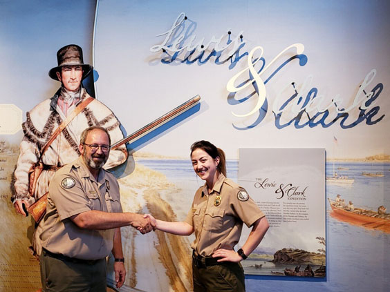 A man dressed in a park ranger uniform shakes a woman's hand in the same attire. Behind them is a mural of Lewis & Clark