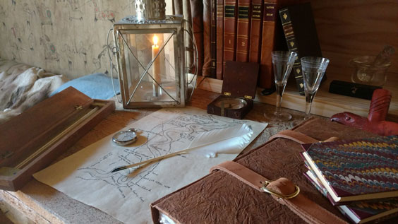 A desk sits scattered with a map, candle, glasses, portfolio, quill, books, and other items