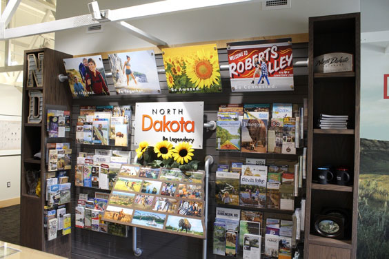 A wooden wall display with many brochures. In the middle is a sign that says North Dakota Legendary, and there are fake sunflowers below it.