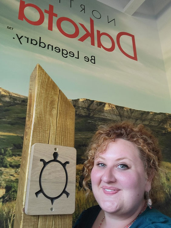 A woman with her red hair pinned back stands in front of a banner that says North Dakota Be Legendary and has a wooden post with a turtle outline on it.