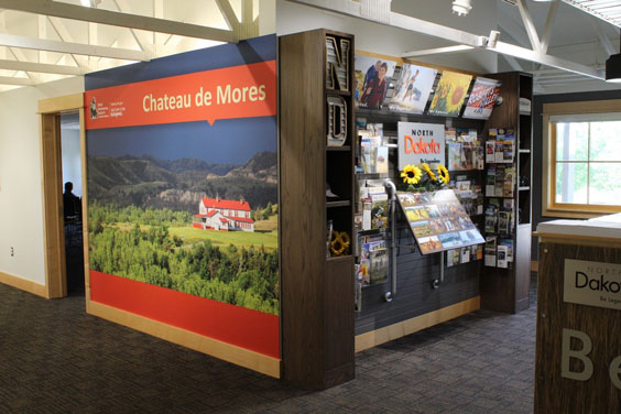 On one  wall is a banner that says Chateau de Mores and has a photo of a qwhite house with red roof surrounded by green grass, trees, and hills. On the other  wall is a display with many brochures.