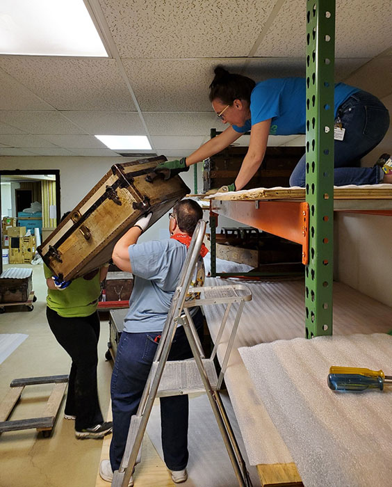 A woman is kneeling on the top shelf of a shelving unit while two other women hand her a large trunk