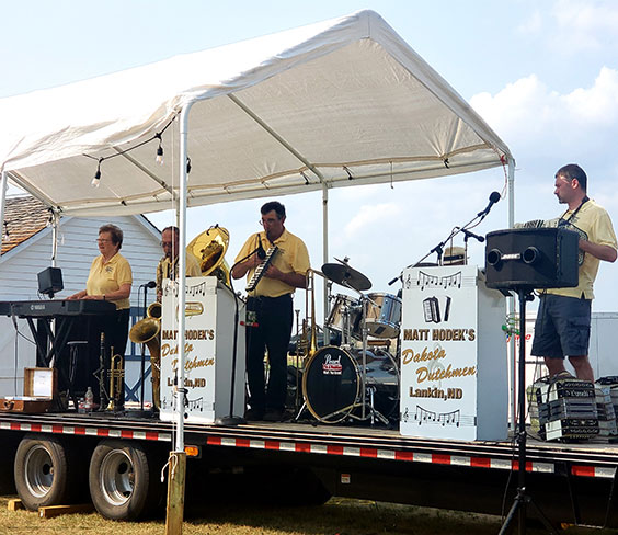 A five person band stands palying on a trailer bed. A woman plays piano while men play sax, keyboard, drums, and accordion. They are all dressed in yellow polo shirts and dark bottoms. Two white podiums sit on the stage and read Matt Hodek's Dakota Dutchmen - Lankin, ND.