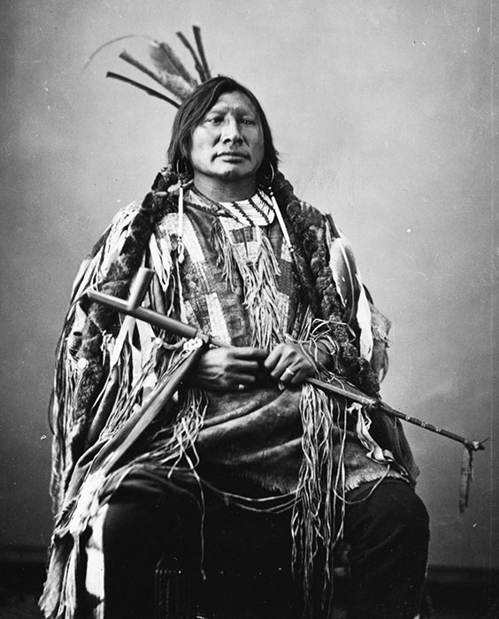 A native man sits wearing traditional attire with long, braided hair and a peace pipe in his hands