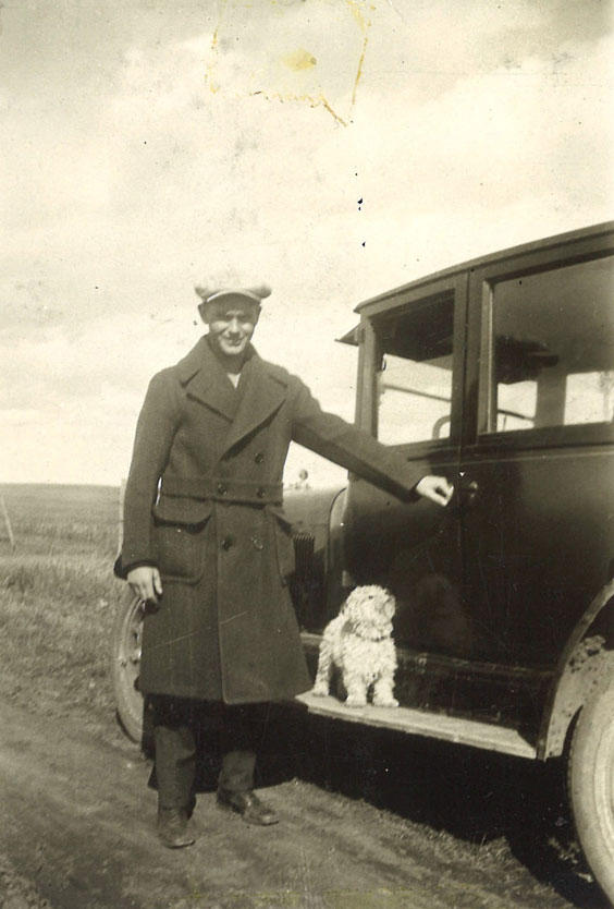 A man in a long trenchcoat and hat stands next to a dark colored car with a white dog on the running board