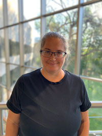 A woman wearing a black tshirt and black framed glasses with her hair pulled back stands in front of a window with trees in the background