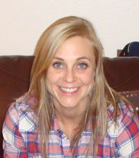 A young blonde woman wearing a red, white, and blue plaid shirts sits on a brown chair smiling at the camera