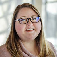 A young woman with long blonde hair and dark framed glasses smiles at the camera