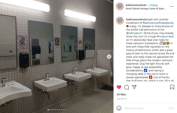 A public bathroom is pictured with four sinks with mirrors above each with square tiles on the walls and historical posters between each mirror