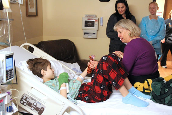 An adult woman sits on a hospital bed where a young boy lays with his hand wrapped. She is handing the woman is handing the boy a small treat bag.